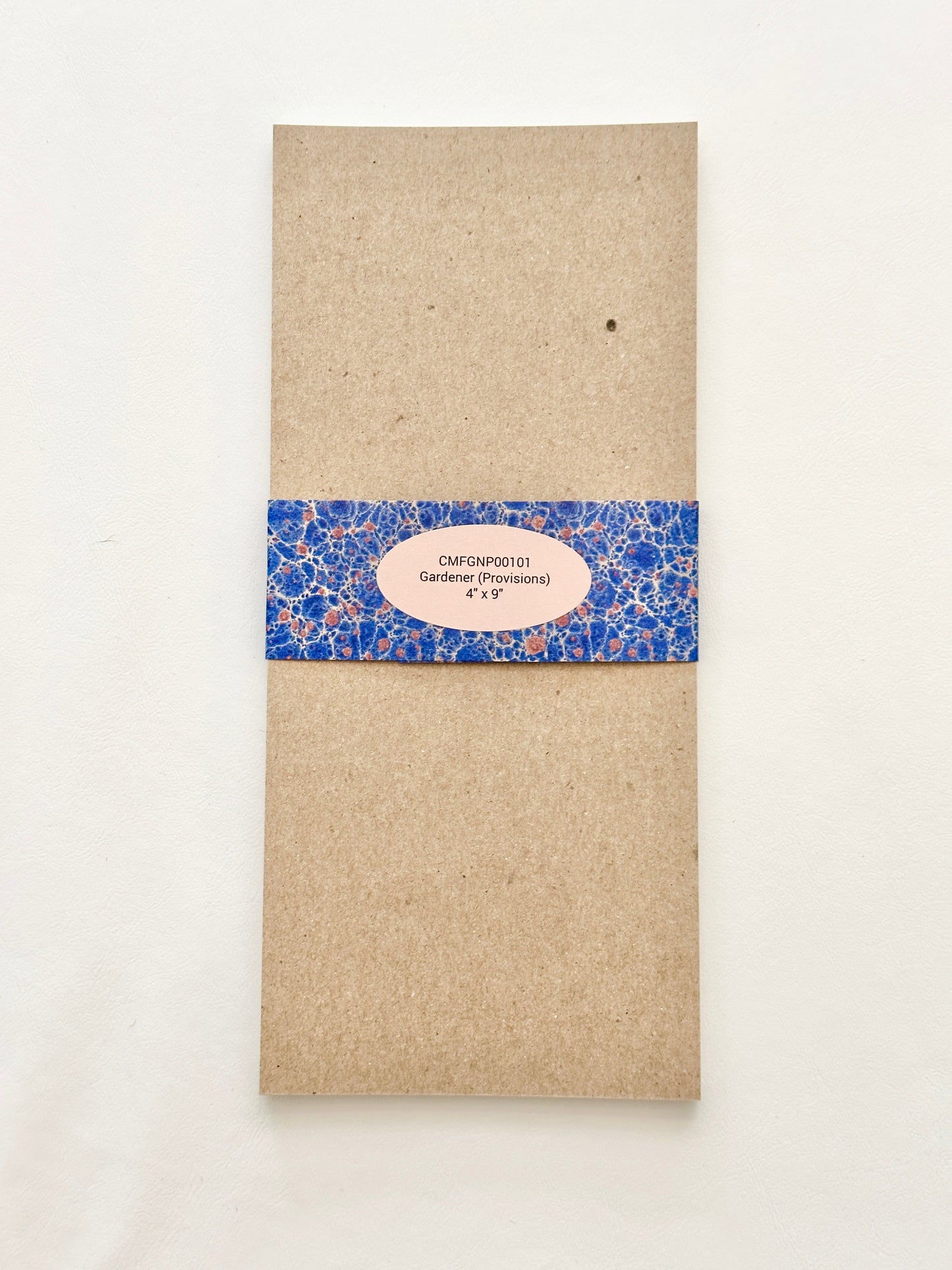 Brown chipboard back of notepad wrapped in a blue patterned bellyband with a pink oval label in the center.