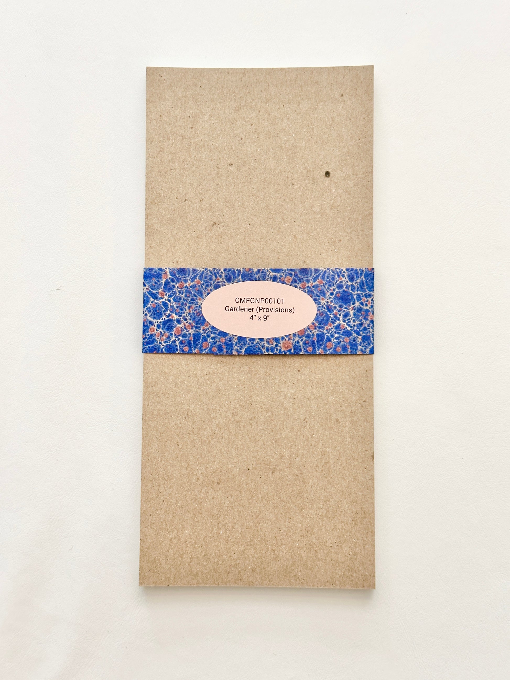 Brown chipboard back of notepad wrapped in a blue patterned bellyband with a pink oval label in the center.